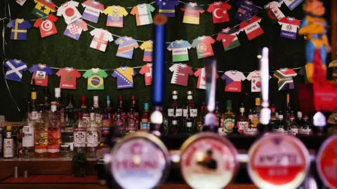 AndyRain/EPA/Shutterstock A bar with bottles of drink on and a decoration of small shirts from different teams in the World Cup. 