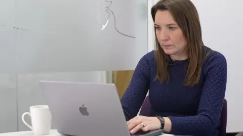 JAMIE NIBLOCK/BBC Nikki Fox sits at a desk wearing a deep purple woollen dress. There is a frosted glass wall to her right behind her and a white cup to her right on a white desk. Her silver laptop sits in front of her, while a black phone is on the large desk to her left. She is looking at a screen and concentrating on the conversation.