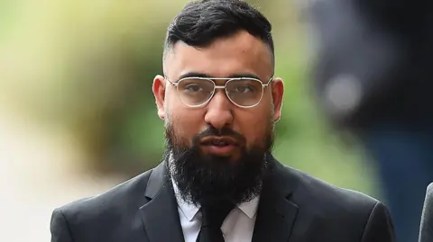 Muhammed Amaad standing outside court in a black suit wearing silver-rimmed glasses and a black suit with a white shirt and black tie. 