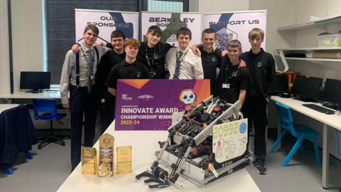 Eight Berkeley Green UTC pupils stand together in a classroom between their robot on the table