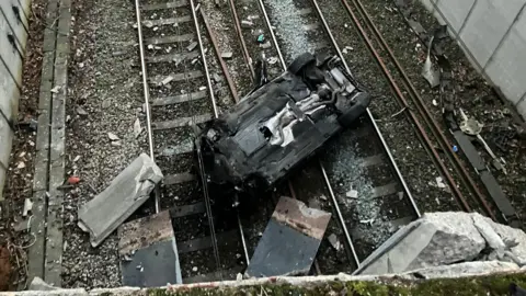 A burnt-out and smashed BMW lies on its roof on a railway line. Debris is scattered all around the wreckage.