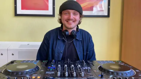 BBC Felix Coulton at the decks. He is wearing a beanie hat and is smiling for the camera.