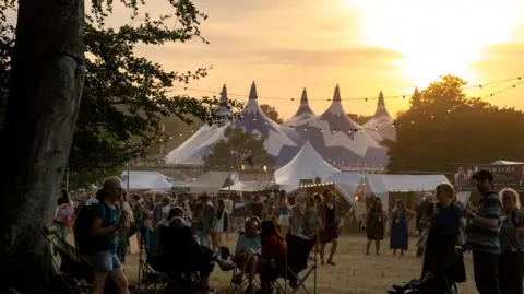 Garry Jones Photography Lots of people move through the festival site at dusk. There is a glowing orange sunset above a large blue and white tent with a pointed roof. Groups of people are sitting in camp chairs and walking through the area, with lots of trees and strings of lights around. 