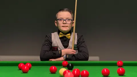 A young boy with glasses is wearing a black shirt, yellow bow ties and tweed waistcoat as he holds a snooker cue with his arms crossed on a snooker table with scattered balls