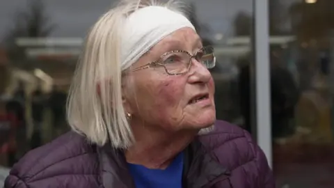 A woman wearing a headband and purple jacket can be seen speaking to someone who is not on camera