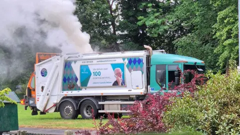 A side view of a white bin lorry, with a turquoise cab. White smoke is billowing from the top of the lorry 