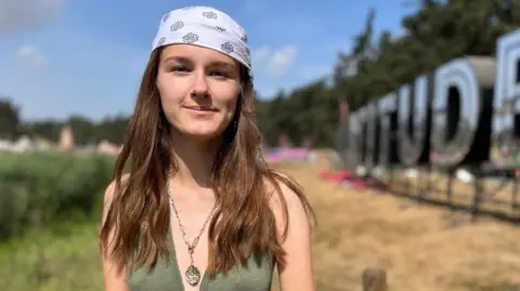 Richard Haugh/BBC A girl is wearing a green top and white bandana. She is in front of a large Latitude sign.
