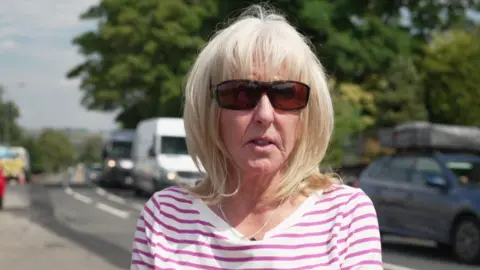 A woman wearing a pink and white striped top and sunglasses stands next to a road looking into the camera with a neutral expression.