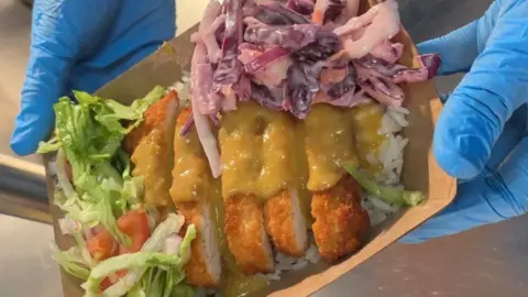 supplied slices of breaded chicken and sauce, in a box with sauce on top and two hands with blue gloves holding up the food.