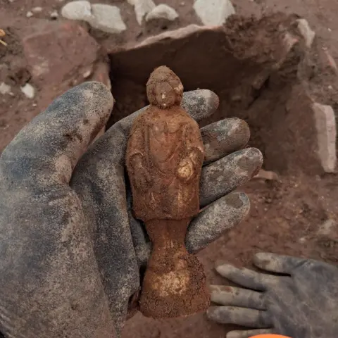 Oxford Archaeology Brown figurine held in black gloves above grave.