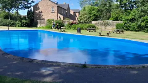 North Yorkshire Council A large blue paddling pool set in parkland. A pavement surrounds one side, and grass on the other, with picnic benches.