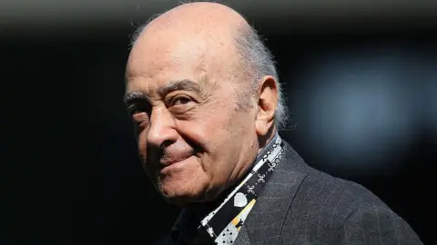 Mohamed Al Fayed is pictured at a football match wearing a grey suit jacket and a patterned black, grey and yellow shirt. He is balding with grey hair to the sides. At the time he was the Fulham chairman and is pictured ahead of the Barclays Premier League match between Fulham and Wolverhampton Wanderers at Craven Cottage on April 17, 2010 in London.