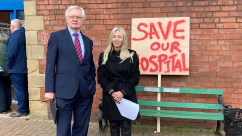 David Davis, dressed in a blue navy suit, stands to the left in the picture.  Michaela wears a black knee-length coat.  Both hold clip boards and papers and stand in front of plackard that says in red writing "Save our hospital".