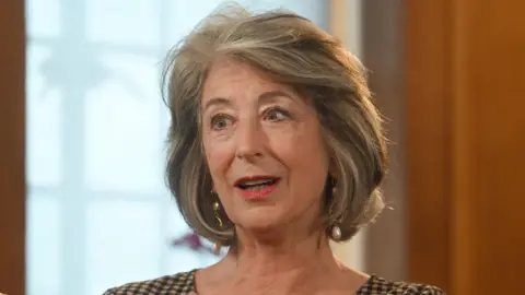 PA Media Maureen Lipman is looking to the side of the camera in mid conversation. She has a bouncy grey bob and is wearing a checked top or dress and and dangly earrings