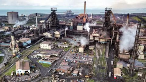 An aerial shot of Tata Steel's Port Talbot steelworks in south Wales. Smoke is coming out of a number of chimneys across the site, while cars are parked in the middle. To the right-hand side rail tracks lead out the works, while the sea can be seen behind it. 