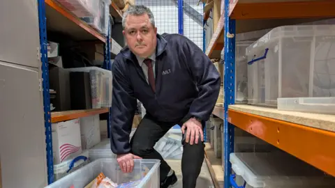 Head of England's Illegal Money Lending Team David Benbow is crouched down over a box, surrounded by shelves, in the evidence store of a warehouse.