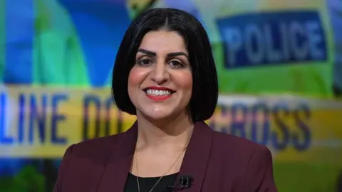 Head and shoulders image of Home Secretary Shabana Mahmood on the set of Sunday with Laura Kuenssberg. She has dark hair in a bob with a centre parting and is smiling at the camera. She is wearing a burgundy blazer over a black blouse and the background shows a graphic of blurred-out police signage