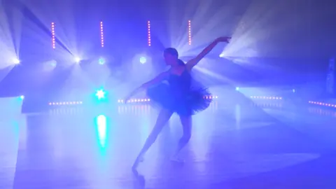 Supplied Jemima performing on stage. Behind her is purple lights and she is wearing a tutu. 