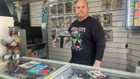 Simon Thake/BBC A bald man in a black sweater stands behind the counter at a shop. The glass cabinet below him contains brightly coloured toys.