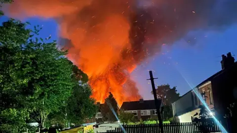 Urooj Imran A picture of the fire with smoke and sparks billowing into the night sky. There are houses and trees in front of it.