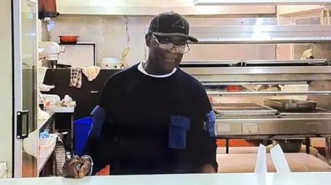 Family handout Mr Salmon is wearing a black cap and black top. He is working in the kitchen of the takeaway.