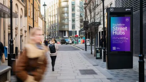 BT Stock photo of a black digital signage unit with a large vertical screen on it standing in an urban landscape. The screen, in purple and blue, reads "Say hello to Street Hubs" with a BT logo in the corner. Two women are visible, one walking away from the camera, with another in the foreground blurred by movement.