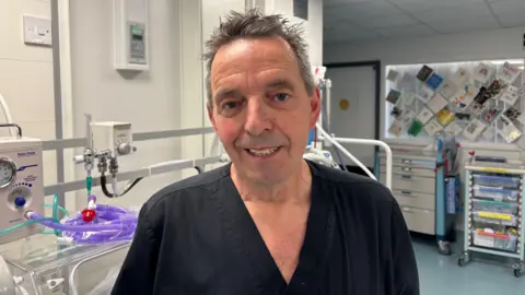 A man with silver hair and wearing black hospital scrubs smiles for the camera