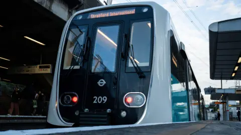 Exterior of DLR train at a platform shows white and green livery with the destination Stratford International in yellow writing