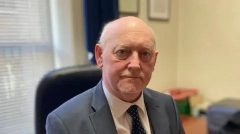A man with short white hair and beard is looking into the camera, unsmiling. He is wearing a grey suit jacket and white shirt with a blue patterned tie. 
