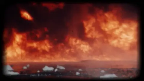 BBC/Wall to Wall Media Archive-style footage shows an intense fire burning across a distant shoreline. Towering flames and smoke dominate the frame, while floating chunks of a white substance drift across the dark water in the foreground.