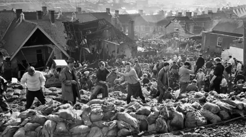 Mirrorpix via Getty Images A black and white image shows rows of rescue workers moving the coal after the Aberfan disaster, with rows of sandbags in front of them. A damaged building can be seen in the background, with the town of Aberfan behind. 