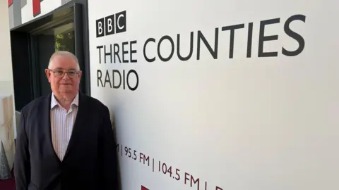 Amy Holmes/BBC Bedfordshire Police and Crime Commissioner John Tizard standing beside BBC Three Counties Radio branding at the station's studios in Dunstable