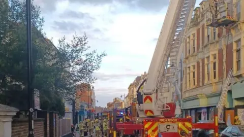 Fire engine with ladder extended up to a four-storey building - numerous firefighters are on the road below and buildings stretch along both sides of the road.