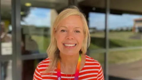 BBC/George Carden A white woman with blonde hair smiling. She is wearing a stripy red and white shirt with a rainbow lanyard. She is facing the camera and stood in front of windows