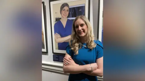 UHCW/The Hunterian Museum A blonde woman in a blue top stands in front of her photographic portrait, in which she is wearing a dark blue surgical top and a head covering over her hair.