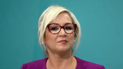 O'Neill has a purple top on and her blonde hair tied back. She is staring at the camera she wears red glasses, makeup and pink lipstick. the background is blue.