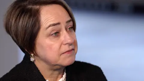 Mandy has short dark hair, is wearing a dark textured jacket and small drop earrings, and is turned slightly to one side with the face in profile.