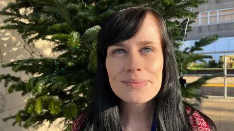 John Devine/BBC Gemma, a woman, with black hair, smiling for the camera. The tree is behind her. She is outside and there is a 1960s concrete building in the background.