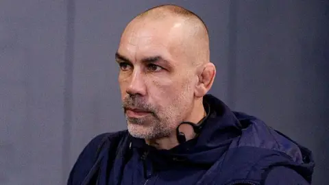 Alex Sanderson looking straight ahead with a grey wall behind him 