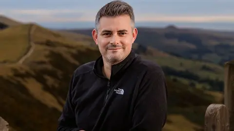 Man with short grey hair black fleece stood infront of hillside backdrop