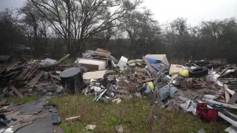 A dense heap of illegally dumped waste in a wooded area, featuring broken wooden boards, plastic bags, tyres, metal scraps and other discarded materials.