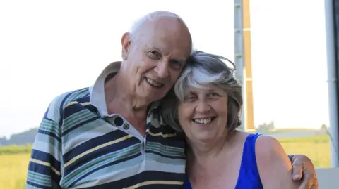 Family handout A man and woman embrace and smile at the camera. The man is bald with some grey hair and he is wearing a white, blue, yellow and green striped polo T-shirt. The woman has short grey hair and is wearing a blue vest top.