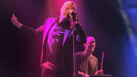 Getty Images Blancmange performing at a gig. Two members of the band are visible, including Neil Arthur, the vocalist. He is standing at the mic and is dancing to the music with his eyes closed. His left hand grips the microphone, while his right is raised. The drummer is behind him and to the right, with one stick raised.