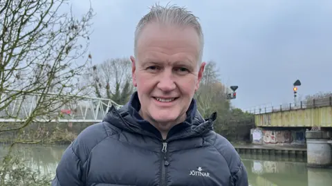 Steve Morgan appearing with grey hair and black puffy jacket smiles at the camera in front of a river, with a railway bridge crossing the river to the right of the image and a white painted metal pedestrian bridge to the left 