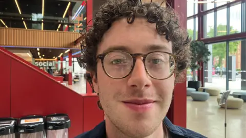 Kieran Minto, pictured from the shoulders up, with short curly brown hair. He is wearing round glasses. 