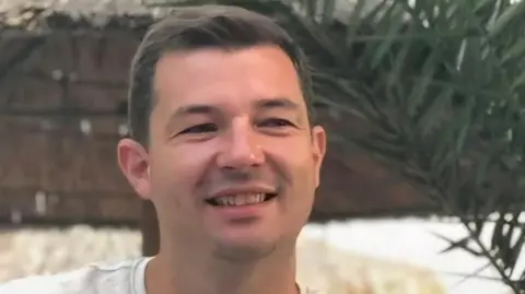 David Lusby A smiling man in a white tshirt sits in front of a brick wall. He has short dark hair