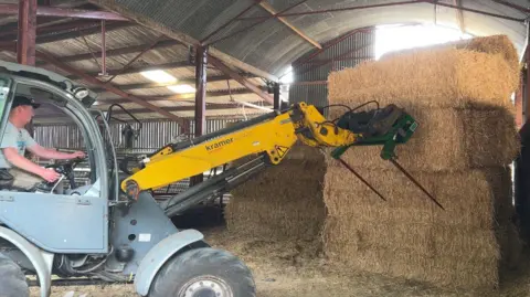 Tom in the cab of a farm machine with a long yellow front lifter on it, attached to two prongs which he will use to pick up bales of hay.