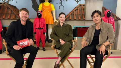 Roman Kemp and two actors from Squid Game smile to camera, while seated in chairs.