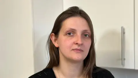 Kate Bradbrook/BBC Lauren has dark hair and brown eyes. She is looking into the camera and standing in front of white kitchen cupboard doors. 