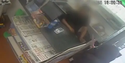 A person who is hooded is holding a knife pointing it at a shopkeeper over a counter